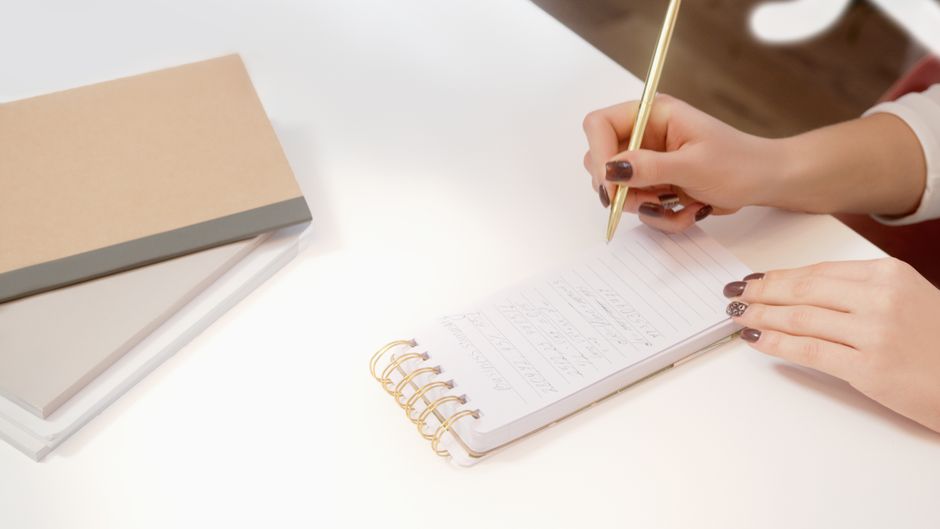 A close-up of hands writing in a spiral notebook on a white desk.