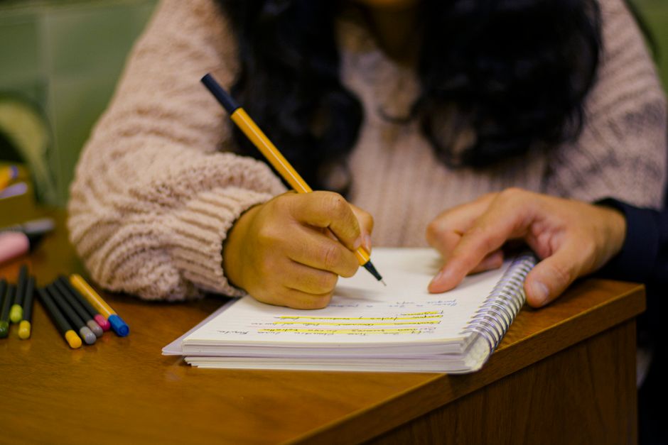 A person writing notes in a notebook with colorful pens on a wooden desk.