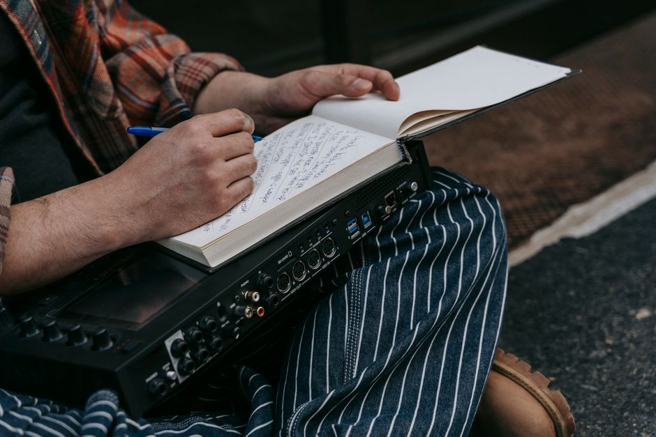 Close-up of a person sitting with a notebook, writing music notes, and holding audio equipment.